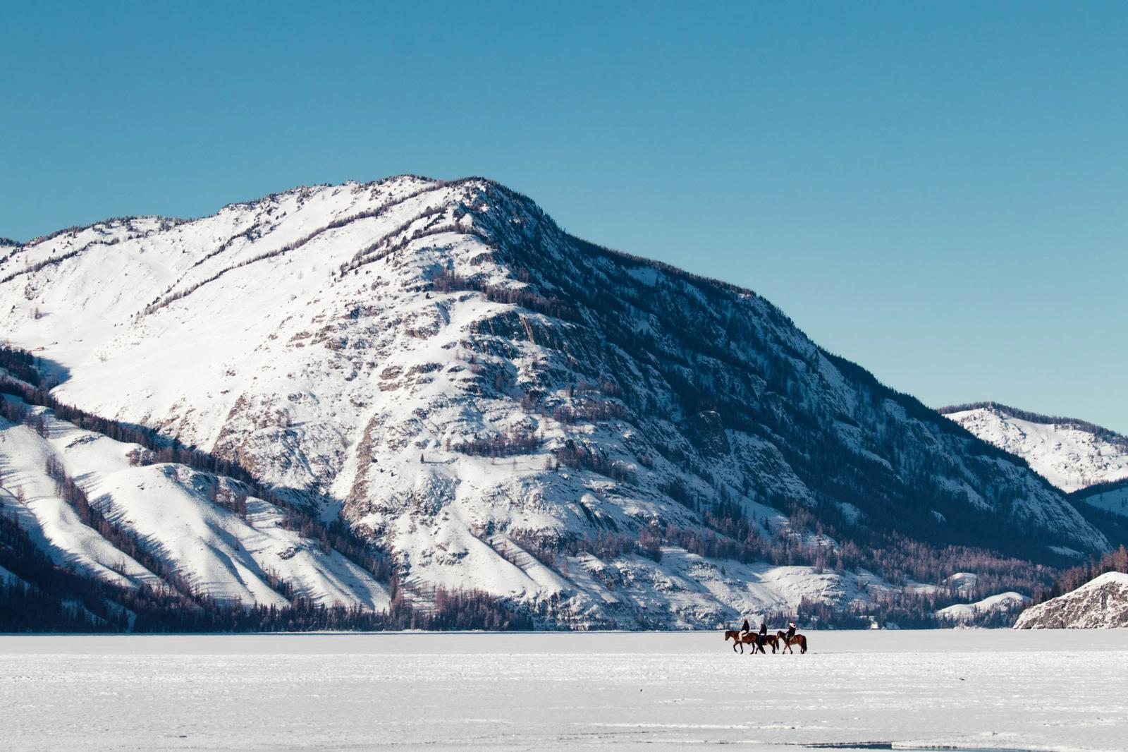 Picture of an expansive view of snow capped mountains and a few people riding horses in a distance