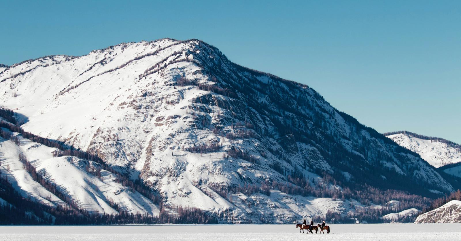 Picture of an expansive view of snow capped mountains and a few people riding horses in a distance